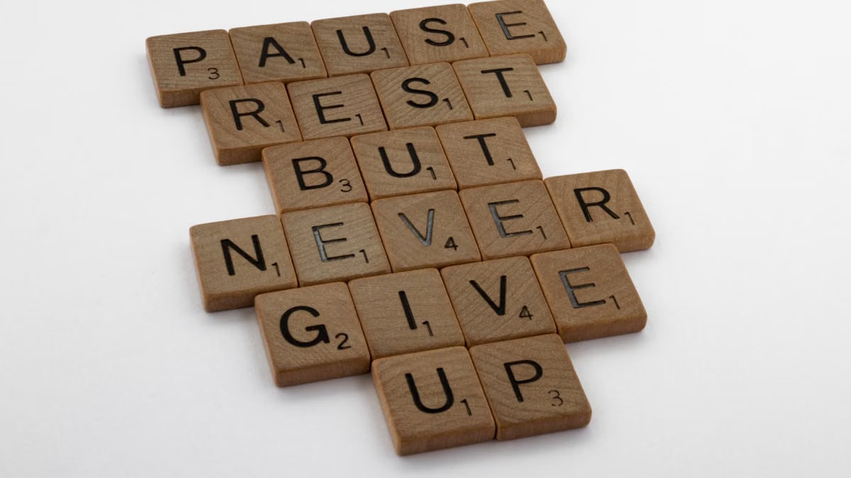 Wooden tiles spelling out 'Pause Rest But Never Give Up' on a white background.